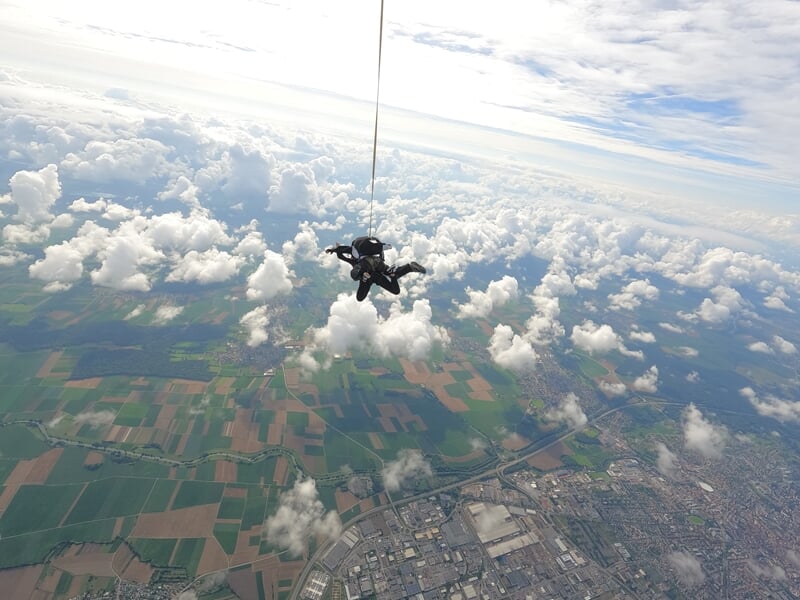 Parachutiste en solo vu de haut, survolant une campagne avec champs et nuages sous un ciel partiellement couvert.
