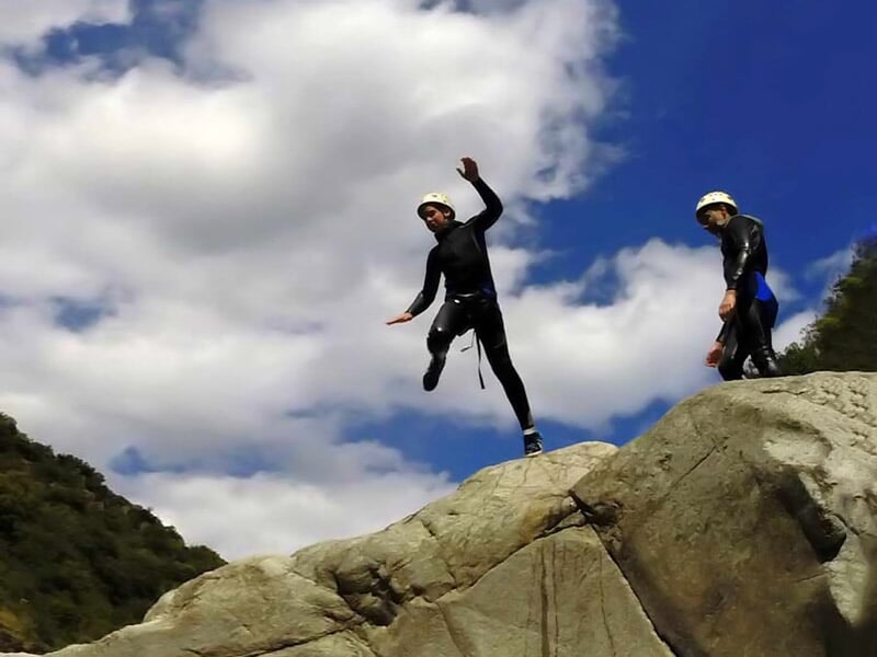 Personne en combinaison noire et casque sautant d'un rocher dans un paysage naturel avec ciel bleu et nuages.