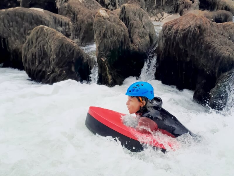 Personne en casque bleu et combinaison nageant avec un flotteur rouge dans une eau agitée près de rochers couverts d'algues.