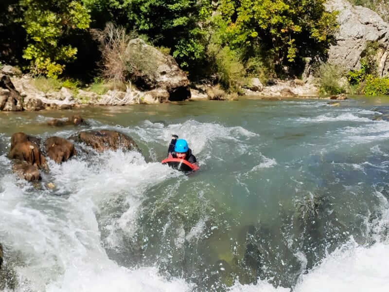 Participant en casque bleu et combinaison nageant dans une eau vive avec des remous et des rochers visibles.