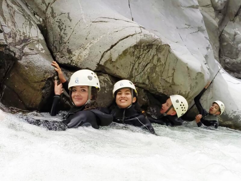Quatre personnes souriantes en combinaison et casque tenant les rochers dans un courant d'eau vive en canyoning.