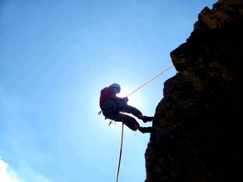 Un grimpeur en rappel descend une paroi rocheuse, silhouetté contre le ciel avec le soleil en arrière-plan.