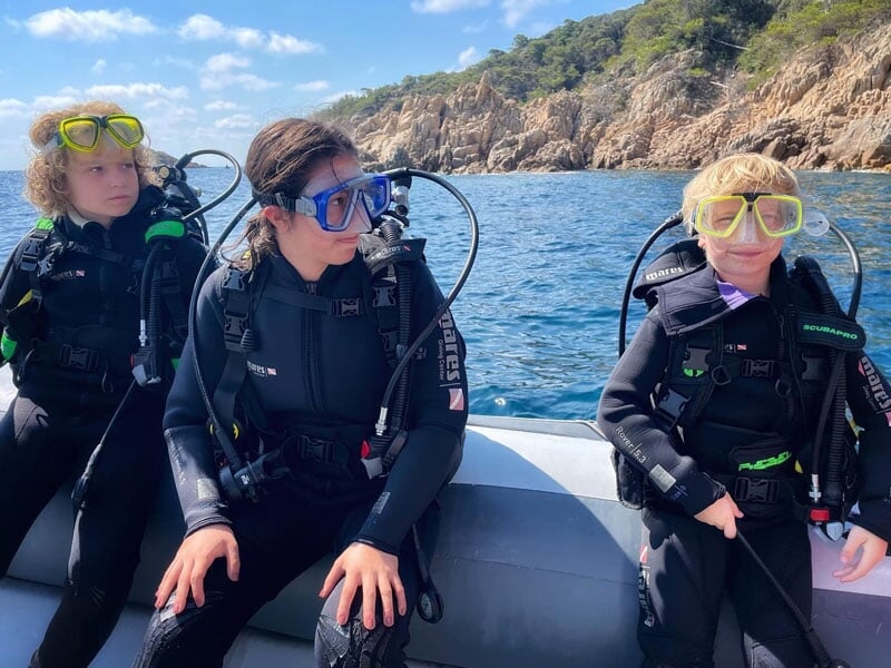 3 enfants sont assis sur le bord du bateau en combinaison de plongée