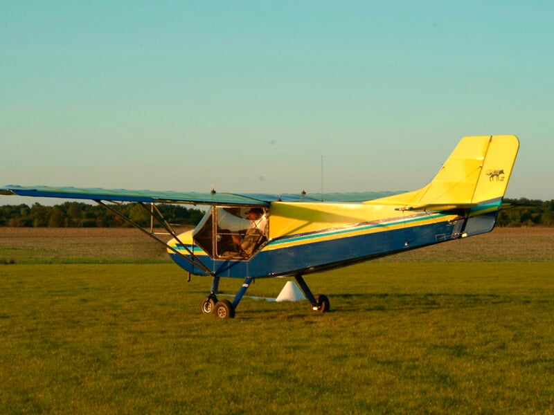Petit avion ultraléger jaune et bleu posé sur une prairie verte sous un ciel clair au coucher du soleil.