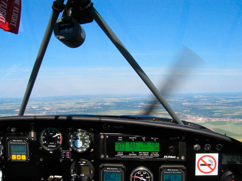 Vue depuis le cockpit d'un avion ultraléger en vol, avec instruments de bord et paysage de campagne en arrière-plan.