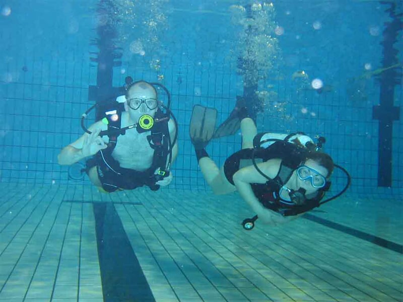 Deux personnes en plongée sous-marine dans une piscine, équipées de masques, détendeurs et combinaisons de plongée.