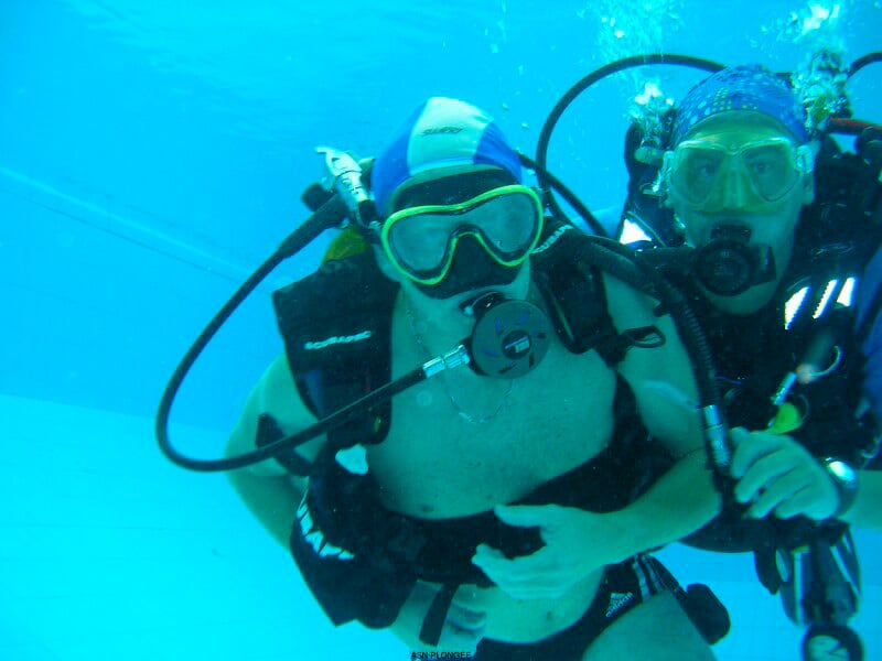 Deux plongeurs en scaphandre complet sous l'eau dans une piscine, l'un guidant l'autre pour un baptême de plongée.