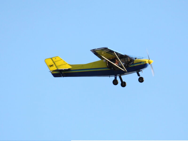 Avion ultraléger jaune et bleu en vol dans un ciel bleu clair, vu de côté avec hélice en mouvement.