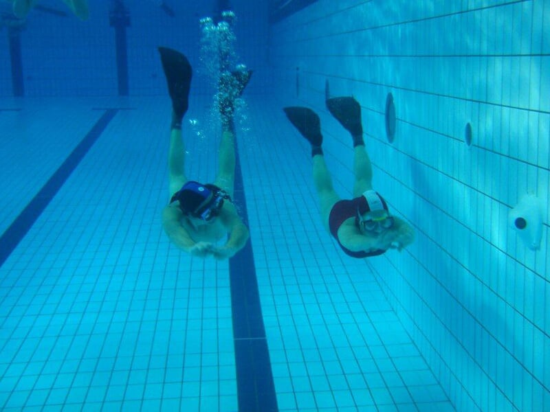 Deux plongeurs sous l'eau dans une piscine, nageant côte à côte avec palmes et masque, près du fond carrelé.