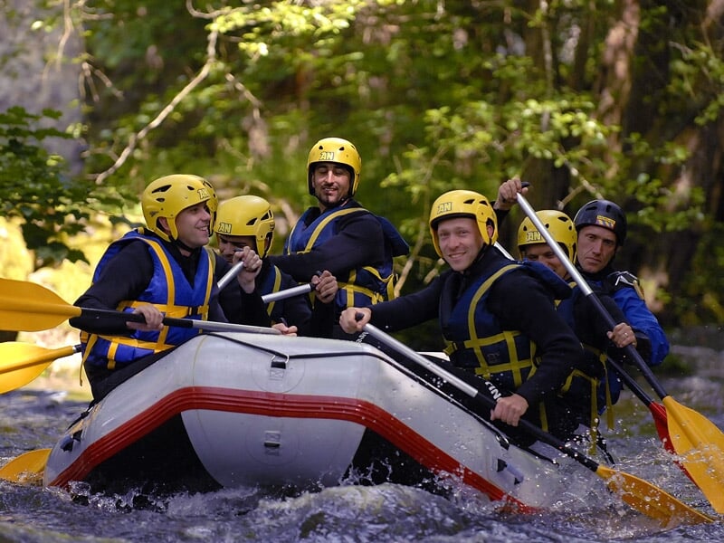 Six personnes en raft bleu pagaient dans une rivière avec des rapides sous un ciel clair.