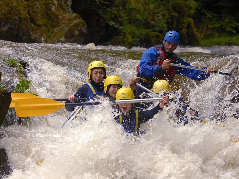 Un groupe de six personnes en raft bleu pagaient dans une rivière avec des rapides et rochers.