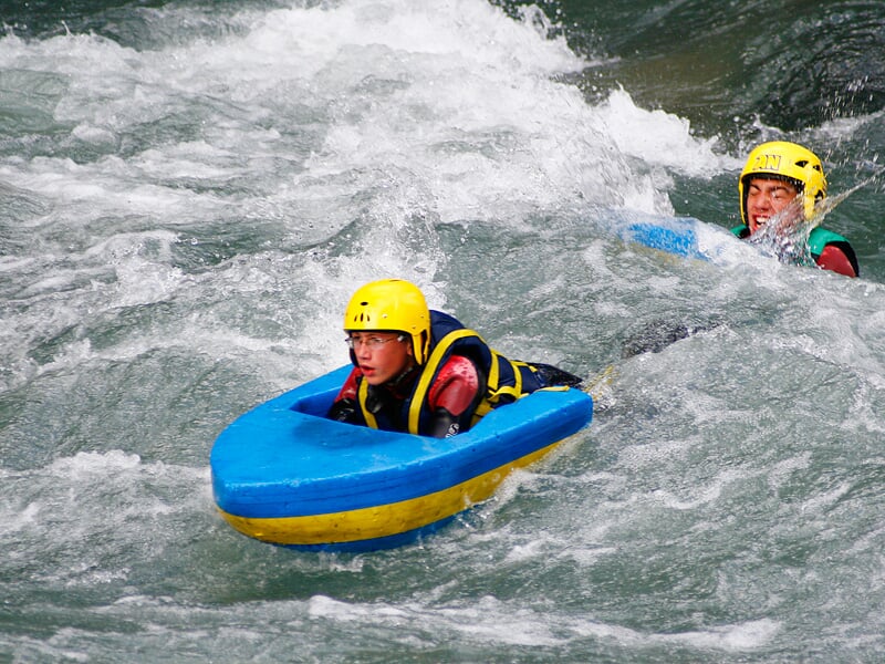 Un jeune homme en hydrospeed descend une rivière tumultueuse avec concentration et casque jaune.