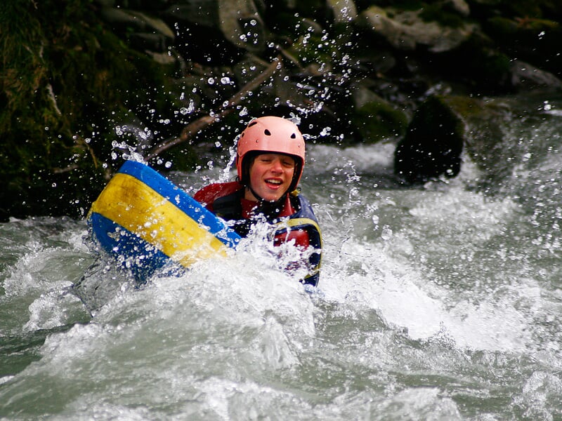 Un homme souriant en hydrospeed affronte les rapides d'une rivière entourée de végétation dense.
