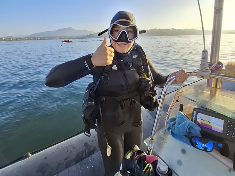 Plongeuse souriante sur un bateau, portant une combinaison et un masque de plongée, levant le pouce en signe d'approbation.