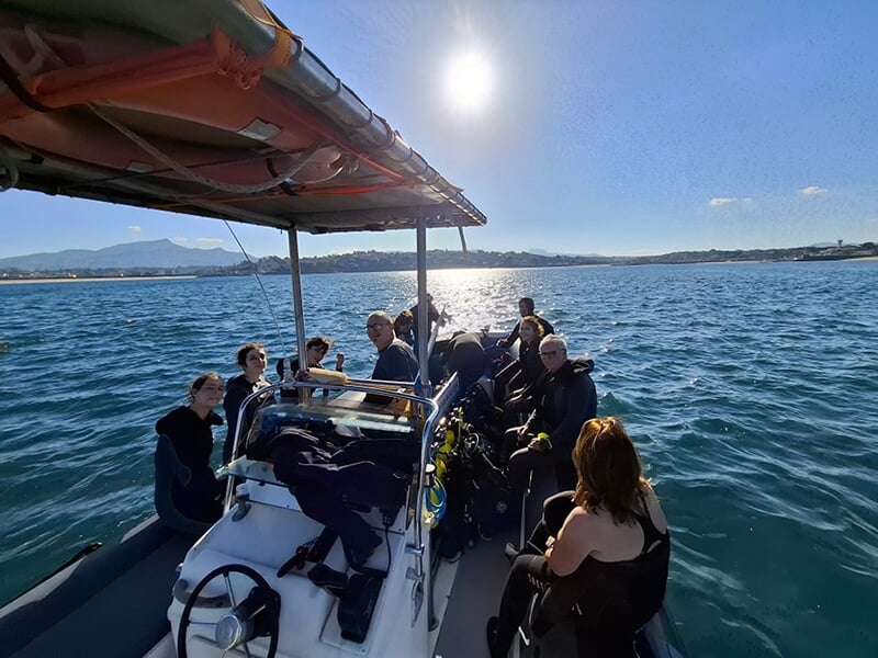 Groupe de plongeurs en combinaison sur un bateau en mer, avec un ciel ensoleillé et des montagnes en arrière-plan.