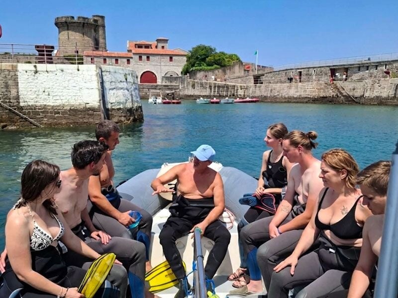 Groupe de personnes en combinaison de plongée assises dans un bateau gonflable près d'un port avec un château en arrière-plan.