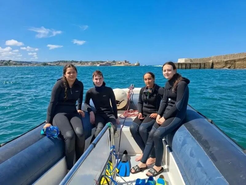 Quatre jeunes en combinaison de plongée assis sur un bateau gonflable avec l'océan et un port en arrière-plan.