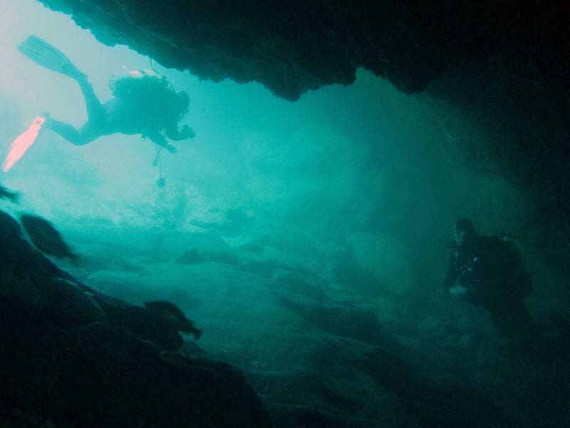 Deux plongeurs sous l'eau nageant près d'une grotte sous-marine avec une lumière bleue diffuse.